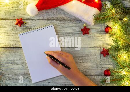 Hand der Frau Schreiben Brief auf Notizblock Papier mit Weihnachten Dekorationen und Bokeh auf weißem Holzhintergrund Stockfoto