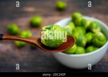 Ein Holzlöffel mit einem frischen Rosenkohl. Weiße Schale mit Rosenkohl im verschwommenen Hintergrund. Stockfoto