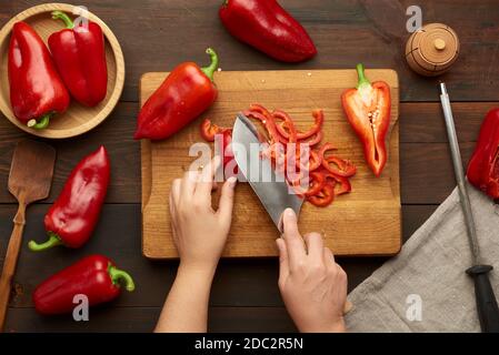 Frau schneidet roten Paprika in Stücke auf Holzbrett, braun Holztisch, Draufsicht Stockfoto