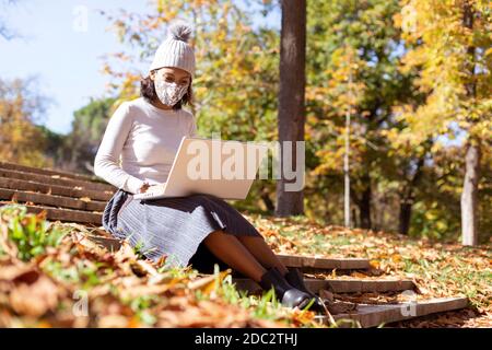 Brunette Frau arbeitet mit ihrem Laptop im Freien. Sie setzt sich und trägt eine Gesichtsmaske. Leerzeichen für Text. Stockfoto