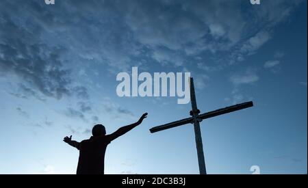 Mann mit ausgestreckten Armen, der unter dem Kreuz auf dem Gipfel des Berges betet. Stockfoto