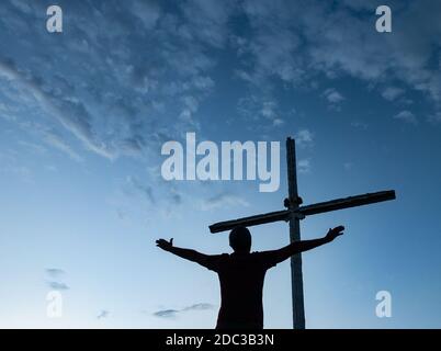 Mann mit ausgestreckten Armen, der unter dem Kreuz auf dem Gipfel des Berges betet. Stockfoto