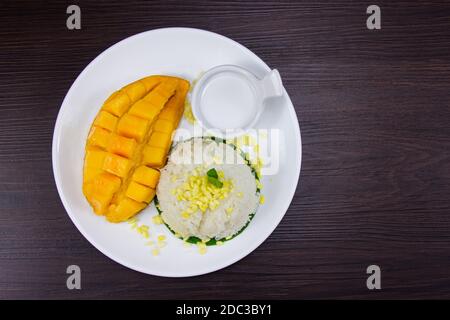Klebriger Reis mit frischer, reifer Mango mit Kokosmilch.die weiße Platte auf den braunen dunklen Holztisch legen ist ein tropisches Dessert im Thai-Stil. Draufsicht. Stockfoto