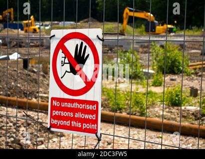 Warnschild an einem Drahtzaun, der eine Baustelle mit einem Bagger und Fundamenten im Hintergrund schützt. Stockfoto