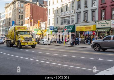 Geschäfte und Geschäfte entlang einer belebten Straße in Chinatown, Manhattan. Chinesischer Stil und Kultur in der Nachbarschaft. Ein Yellow Cement Mixer Truck fährt vorbei Stockfoto