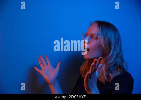 Junge kaukasische Mädchen in Brille schaut in Überraschung auf einem blauen Neon Hintergrund. Seitenansicht und Kopierbereich. Emotionskonzept. Stockfoto