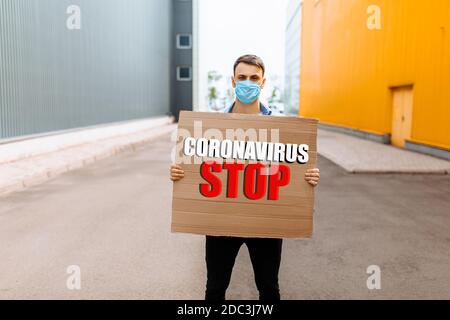 Mann in einer medizinischen Schutzmaske hält ein Pappplakat mit der Aufschrift Stoppt das Coronavirus, während ihr in der Stadt steht, Stoppt Stockfoto