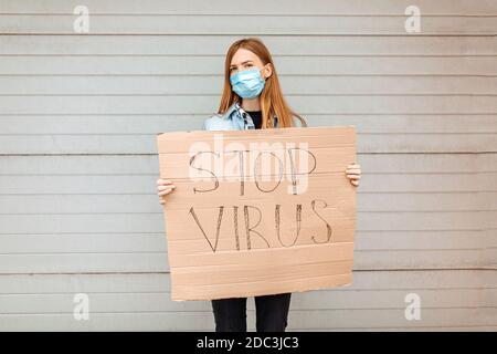 Eine junge Frau in einer medizinischen Schutzmaske hält ein Pappplakette mit der Aufschrift Stop the Coronavirus, steht in der Stadt gegen eine graue Wand. Quarantäne Stockfoto