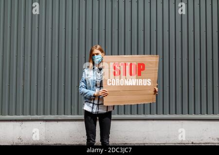 Eine junge Frau in einer medizinischen Schutzmaske hält ein Pappplakette mit der Aufschrift Stop the Coronavirus, steht in der Stadt gegen eine graue Wand. Quarantäne Stockfoto