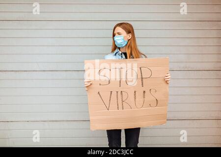 Eine junge Frau in einer medizinischen Schutzmaske hält ein Pappplakette mit der Aufschrift Stop the Coronavirus, steht in der Stadt gegen eine graue Wand. Quarantäne Stockfoto