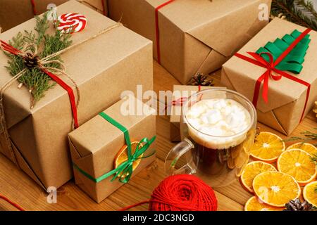 Gruppe von weihnachtsgeschenkschachteln in Kraftpapier eingewickelt, gebunden mit Garn und Tasse Kaffee mit Marshmallow auf Holztisch. Stockfoto