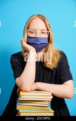 Kaukasische Studentin in einer Maske sitzt mit einem großen Berg von Büchern. Isolations- und Online-Lernkonzept während des Coronavirus. Stockfoto