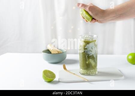 Frau Hand quetscht Limette in Glas mit Eis grünen Matcha Latte auf weißem Hintergrund. Iced Matcha Limonade. Stockfoto