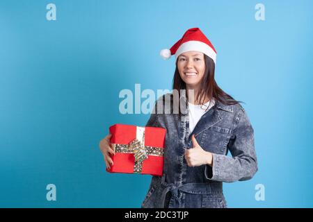 Mock up von glücklich asiatische junge Frau in Santa Claus hut hält eine rote Geschenkbox und gibt einen Daumen Oben auf einem blauen Hintergrund Stockfoto