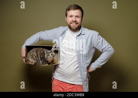 Zufriedener, lächelnder bärtiger Mann hält einen Karton, in dem seine geliebte Katze sitzt und mit Interesse schaut. Grüner Hintergrund. Stockfoto