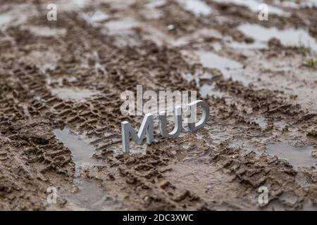 Das Wort Schlamm aus silbernen Metallbuchstaben auf nasser Schmutzoberfläche mit selektivem Fokus, Hintergrundunschärfe und linearer Perspektive in bewölktem Herbsttag li Stockfoto