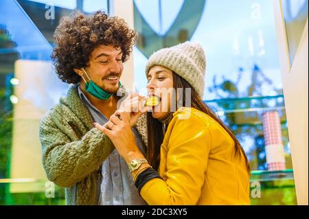 Schön glücklich paar füttern sich gegenseitig in Konditorei - Der junge Mann füttert seiner Freundin einen Makaron - Neues normales Lifestyle-Konzept in covid-19 t Stockfoto