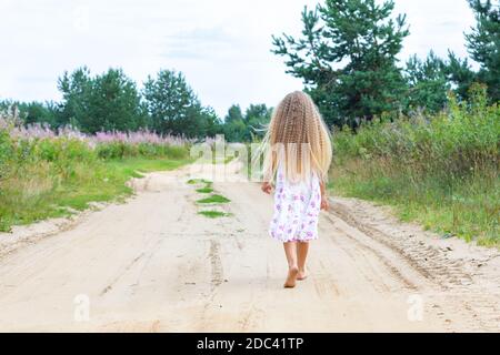 Ein Mädchen mit langen blonden lockigen Haaren geht entlang einer ländlichen Straße. Porträt von hinten in voller Länge. Ein Spaziergang an der frischen Luft in Einsamkeit. Stockfoto