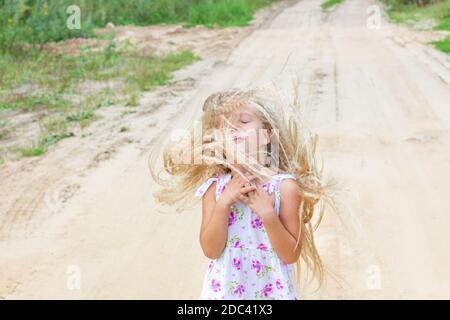 Schönes Mädchen mit üppigen lockigen blonden Haaren schloss ihre Augen, faltete ihre Arme über ihre Brust, schlug ihr Haar, steht auf sandigen Straße. Gefühle, Emotionen. Stockfoto