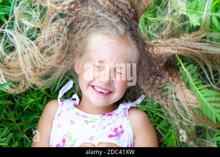 Ein kleines kaukasisches Mädchen mit langen blonden Haaren in einer weißen Sundress liegt auf ihrem Rücken im Gras, schloss die Augen, lächelt, lacht. Kindheit, Glück, Stockfoto