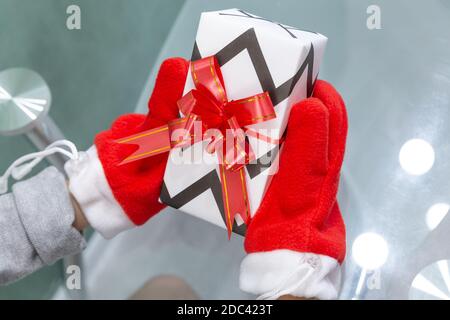 Wunderschön gewickeltes Weihnachtsgeschenk mit einem roten Band mit einer Schleife in den Händen mit roten Fäustlingen des Weihnachtsmannes über einem Glastisch gebunden. Neujahr, Weihnachten, Wint Stockfoto