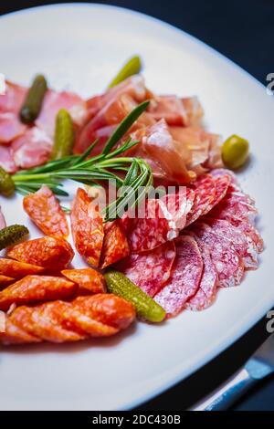 Köstliche Fleischsnacks im Restaurant. Spanischer Jamon in Scheiben, geräucherter Schinken, Pepperoni Würste auf weißem Teller mit eingelegten Gurken, grüner Olive serviert Stockfoto