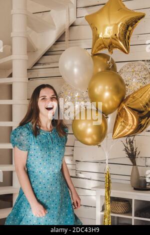 Junge schöne Mädchen in blauem Kleid mit weißen Punkten feiert ihren Geburtstag und genießt die goldenen Ballons. Geburtstag allein zu Hause während des selbst-ist Stockfoto