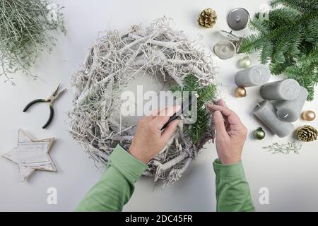 Hände einer Frau, die einen Adventskranz aus hellem Holz mit grünen Tannenzweigen, grauen Kerzen und Weihnachtsschmuck auf einem weißen Tisch bastelt, hoher Winkel Stockfoto