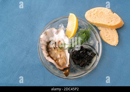 Frisch geöffnete Auster mit Zitrone, Dill und Balsamico-Zwiebeln in Eis in einer Glasschüssel und Brotscheiben auf einer blauen Tischdecke, Kopierraum, Hochwinkelansicht Stockfoto