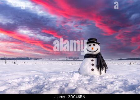 Lustiger Schneemann in stilvollem Hut und schwarzem Skalpell auf schneebedecktem Feld während des Sonnenuntergangs. Epischer Himmel mit lila Wolken auf dem Hintergrund. Frohe Weihnachten und ein glückliches neues Jahr! Stockfoto