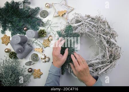 Hände einer älteren Frau, die einen grauen Adventskranz aus Holz mit Tannenzweigen, Lebkuchengebäck, Weihnachtsschmuck und Kerzen auf einem weißen Tabl schmückt Stockfoto