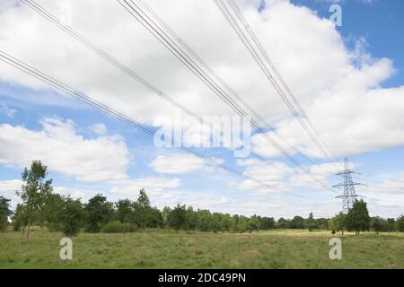 Hell bewölkt weiß und blau Himmel hinter Kopf Stromleitungen und einem entfernten Metall-Strom pylon. Offenes Naturgebiet und Bäume umgeben. Stockfoto