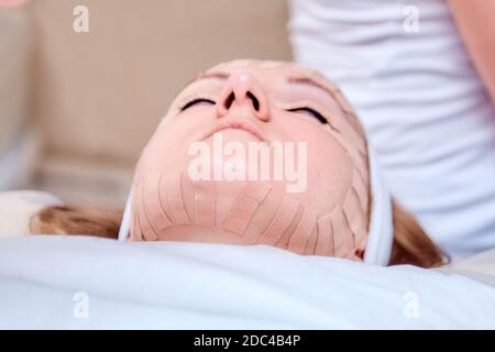 Kinesiotaping. Junge Frau liegt mit Klebeband auf ihrem Gesicht. Physiotherapie und Kosmetologie Verfahren. Methode der nicht-chirurgischen Hautverjüngung. Gesichtshaut Stockfoto