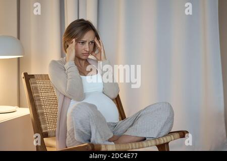 Schwangere Frau, die an Kopfschmerzen leidet, während sie am Tisch sitzt Stockfoto