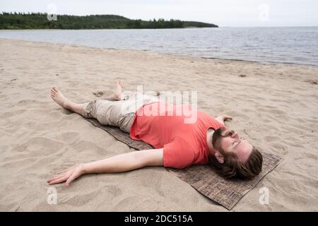 Bärtiger Mann macht Yoga, er macht die Savasana-Übung, entspannt sich in einer Leichenhaltung auf einem Teppich liegend, an einem Sandstrand am See. Stockfoto