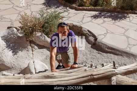 Ein lächelnder junger Mann klettert eine Holzleiter hinauf, die zu einem Höhleneingang an der historischen Stätte des Bandelier National Monument in New Mexico führt. Stockfoto