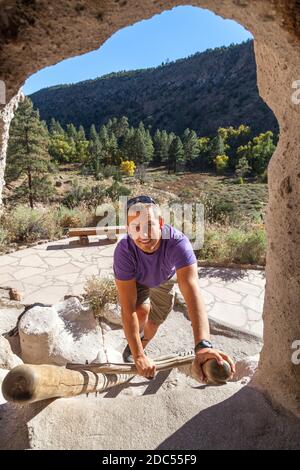 Ein lächelnder junger Mann klettert eine Holzleiter hinauf, die zu einem Höhleneingang an der historischen Stätte des Bandelier National Monument in New Mexico führt. Stockfoto