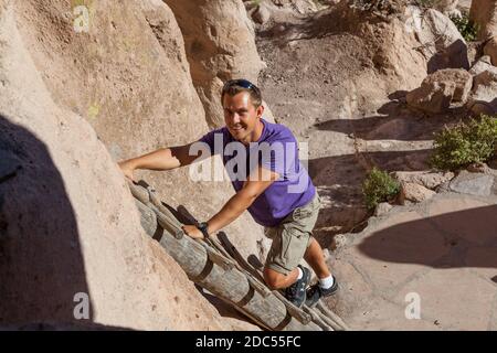 Ein lächelnder junger Mann klettert eine Holzleiter hinauf, die zu einem Höhleneingang an der historischen Stätte des Bandelier National Monument in New Mexico führt. Stockfoto