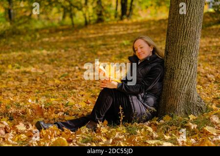 Ein schönes junges blondes Mädchen sitzt auf gefallenen Herbstblättern im Park und lehnt ihren Rücken gegen einen Baumstamm. Das Mädchen hält gelbe Ahornblätter Stockfoto