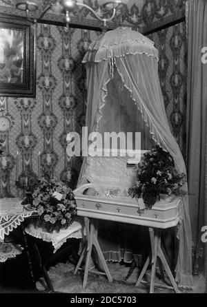 Ein kleines, verstorbenes Baby liegt in einem wunderschön dekorierten, kindergroßen weißen Sarg, um 1920. Der Sarg ist unter einem schicken weißen Baldachin von der Decke abgehängt. Das alles wird in der Ecke des Kinderhauses präsentiert, wahrscheinlich ein Wohnzimmer im Erdgeschoss. Im Sargdeckel ist eine kleine Tafel angebracht, auf der auf Deutsch steht: "Unser Liebling", was "Unser Liebling" bedeutet. Die Schatulle sitzt auf einem Paar spezieller Stativbeine. Um meine Kinder-bezogenen Vintage-Bilder zu sehen, Suche: Prestor vintage Kids Stockfoto