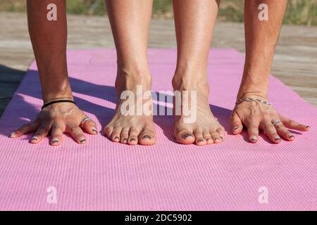 Hände und Füße einer Frau, die einen Yoga macht, strecken sich auf einer rosa Yogamatte auf einer hölzernen Plattform in der Nachmittagssonne. Stockfoto