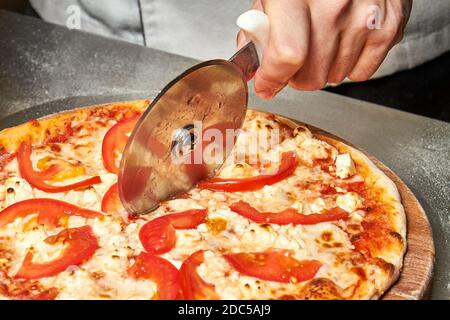 Der Koch schneidet frisch zubereitete Pizza mit Tomaten und Käse Mit einem Spezialmesser Stockfoto