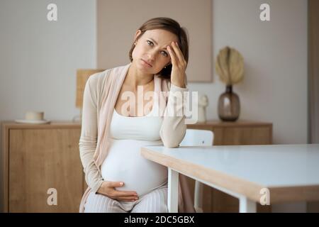 Schwangere Frau, die an Kopfschmerzen leidet, während sie am Tisch sitzt Stockfoto