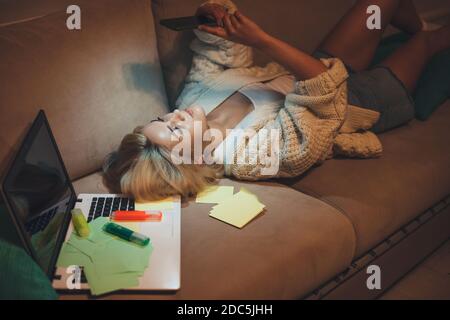 Nachtfoto einer blonden Frau, die auf dem liegt Sofa und Chatten auf dem Handy, während der Arbeit an Hausaufgaben mit Ein Notebook Stockfoto