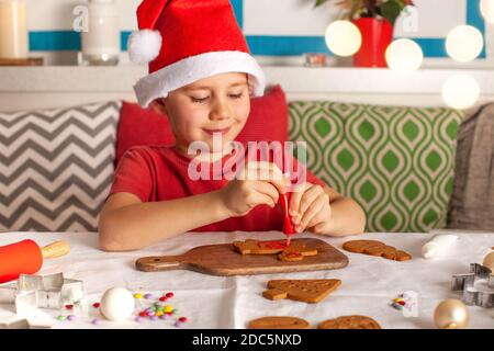 Ein fröhlicher Junge in einem Weihnachtsmannhut schmückt Ingwerkekse in der Küche. Weihnachtsbeleuchtung. Stockfoto