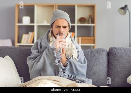 Unglücklicher kranker Mann, der auf der Couch in Decke und gewickelt sitzt Medizin mit Wasser einnehmen Stockfoto