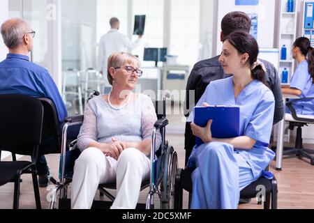Krankenschwester, die Dokumente einreicht, während sie mit behinderten älteren Frauen im Wartebereich des Krankenhauses spricht. Patient fragt nach der Richtung an der Krankenhausaufnahme. Stockfoto
