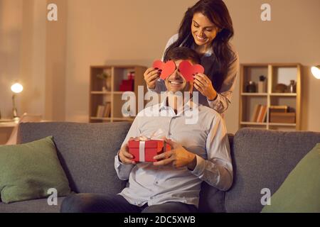 Lächelnde Freundin Abdeckung sitzende Freunde Augen mit Herzen und machen Überraschung zum Valentinstag Stockfoto