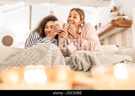 LGBT weibliches Paar, das Spaß unter einer Decke auf der hat Sofa - Cute blonde kaukasische Frau trinken Tasse Tee Im Wohnzimmer lachend mit ihrem schwarzen Hispan Stockfoto