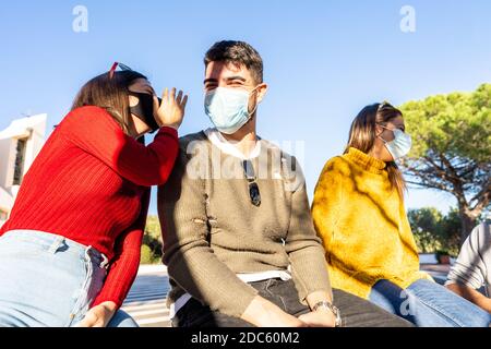 Junge kaukasische Frau spricht im Ohr ihres Freundes Sitzen auf einer Mauer des Stadtplatzes mit Freunden Tragen Coronavirus sicheren Schutz medizinisch Stockfoto